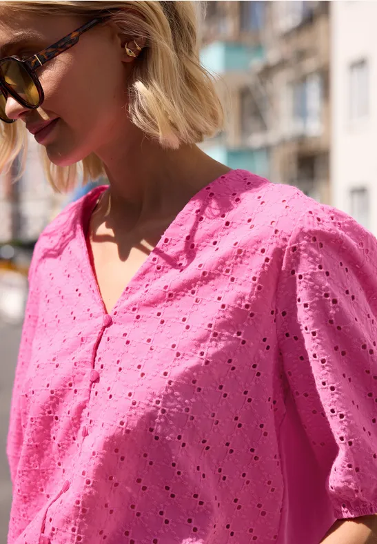 Vrouw met blond bobkapsel draagt roze broderie anglaise blouse met korte mouwen en zonnebril op straat