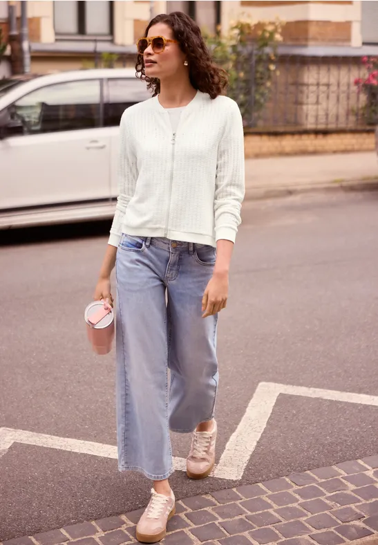 Een vrouw met zonnebril, witte cardigan en jeans, koffiebeker in hand, lopend over straat.