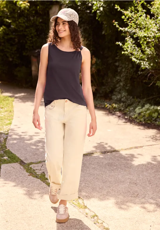 Jonge vrouw loopt op een zonnig pad, draagt zwart hemdje, lichte broek, witte sneakers en een pet.
