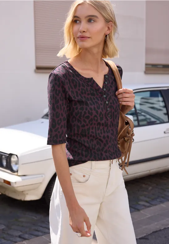 Frau mit kurzem, blondem Haar, gemustertem Top und weißer Hose vor einem weißen Auto.