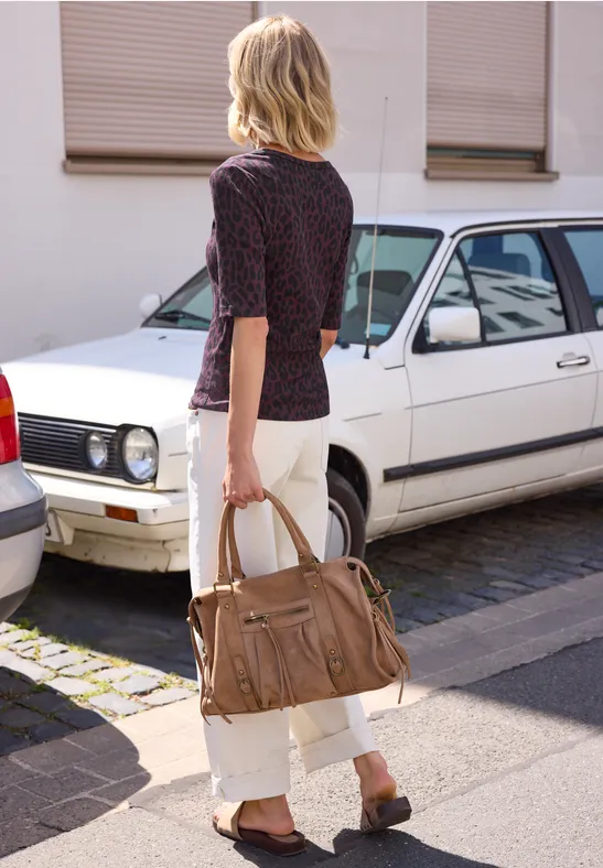 Frau in weißer Hose und gemustertem Oberteil trägt eine braune Handtasche und geht an einem weißen Auto vorbei.