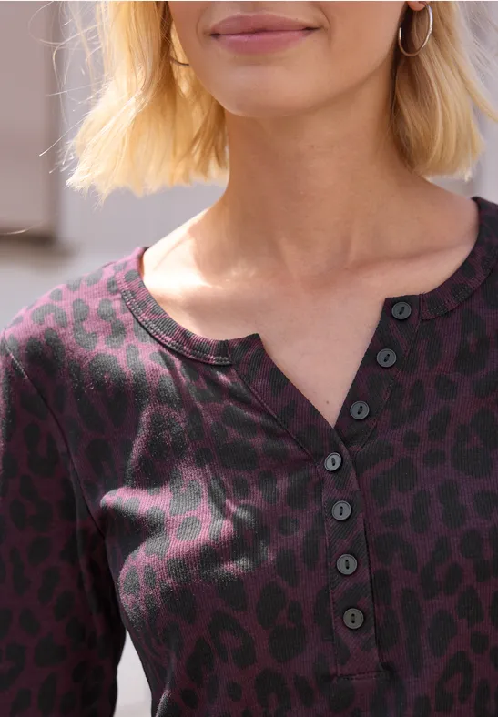 Nahaufnahme einer Frau mit kurzem blonden Haar, die ein lila Shirt mit Leopardenmuster und Knopfleiste trägt.