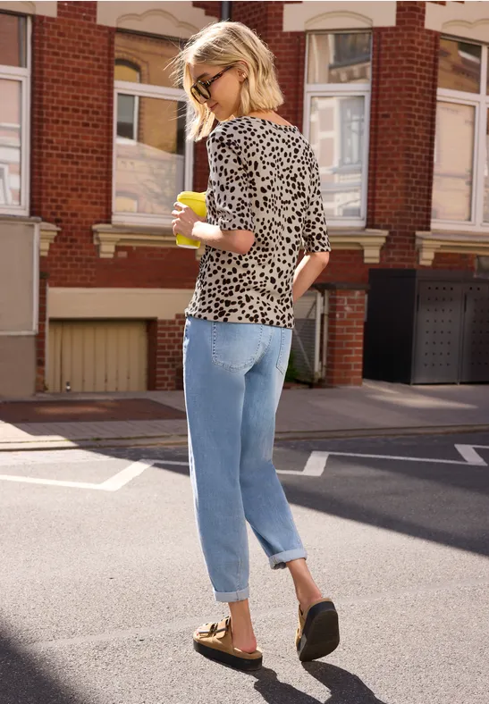 Frau in Leoparden-T-Shirt, Jeans und Loafers schlendert mit Kaffeebecher durch die Stadt.