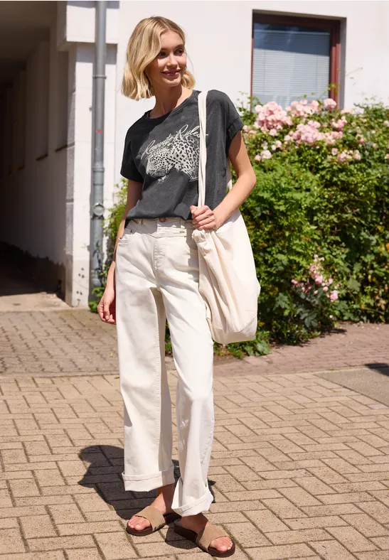 Person in T-Shirt, hellen Hosen und Sandalen auf dem Bürgersteig vor einem Haus mit Rosen trägt weiße Tasche.