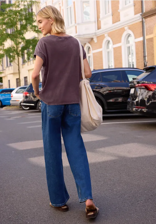 Vrouw in los t-shirt, wijde spijkerbroek en sandalen loopt met stoffen tas langs geparkeerde auto’s in de stad.