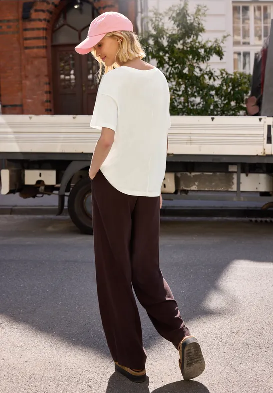 Frau in weißem T-Shirt, Baseballcap und weiten dunklen Hosen spaziert an einem LKW vorbei.