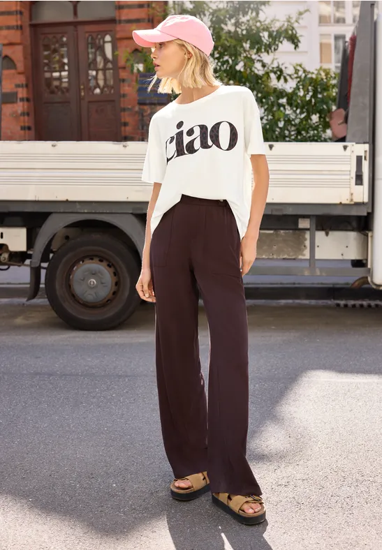 Frau in lockerem Outfit: weißes 'ciao'-Shirt, schwarze Hose, Sandalen und rosa Cap, auf der Straße stehend.