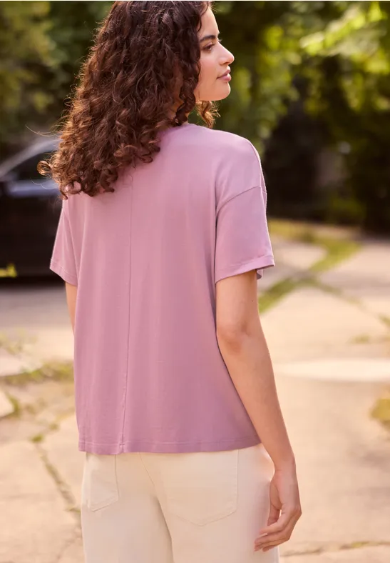Femme vue de dos, cheveux bouclés, t-shirt rose et pantalon clair, debout sur un chemin au milieu de verdure.