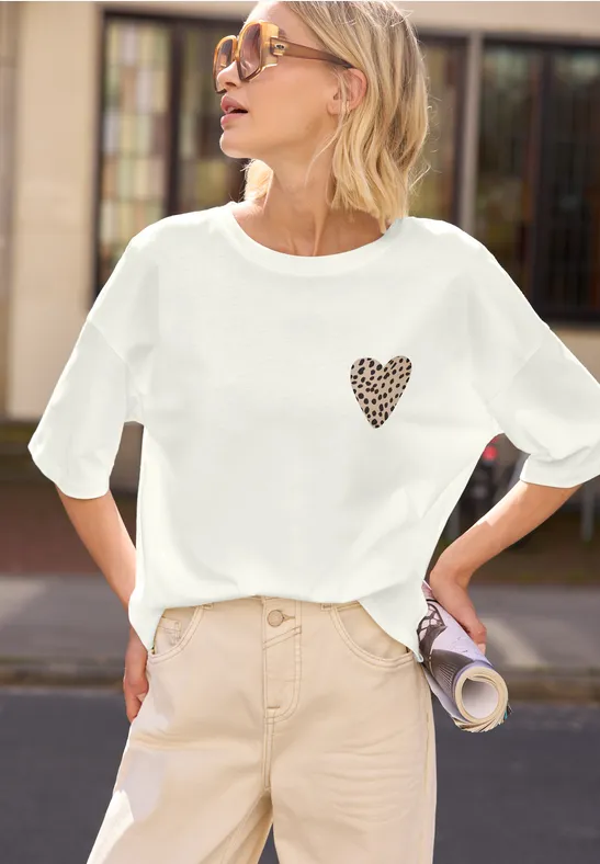 Frau in weißem T-Shirt mit kleinem Herz-Leo-Print, beiger Hose, hält eine zusammengerollte Zeitschrift.
