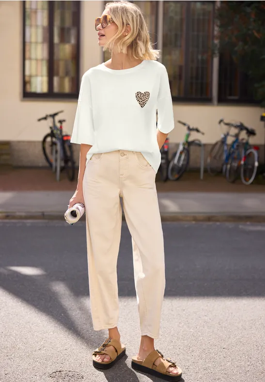 Eine Frau steht auf der Straße, trägt ein weißes Oberteil mit Herzmotiv, beige Hose und braune Sandalen.