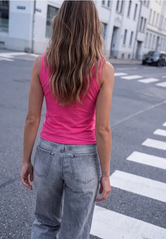 Frau mit langem, gewelltem Haar in pinkem Top und grauen Jeans überquert eine Straße.