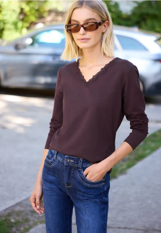 Vrouw met zonnebril, donkerbruine longsleeve en blauwe jeans, staand op de stoep, hand in zak.