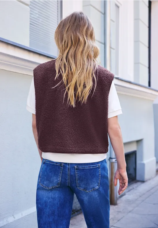 Person von hinten in urbaner Umgebung, trägt braune Weste, weißes T-Shirt und blaue Jeans.