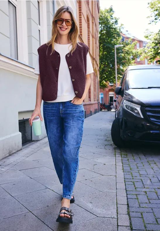 Fröhliche Frau mit Jeans, weißem Shirt, Weste und Kaffee in der Hand geht lächelnd eine gepflasterte Straße entlang.