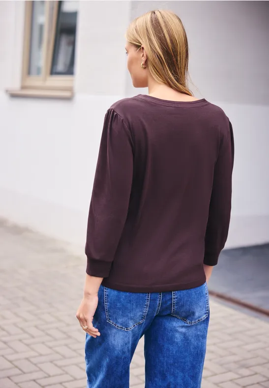 Eine Frau steht mit dem Rücken zur Kamera, trägt dunkles Oberteil und blaue Jeans in urbaner Umgebung.