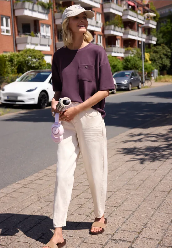 Frau in lila T-Shirt, heller Hose und Cap steht mit Kamera auf dem Gehweg vor Wohnhäusern.
