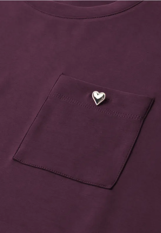 Lila T-Shirt mit Brusttasche und kleinem silbernem Herz-Anstecker.