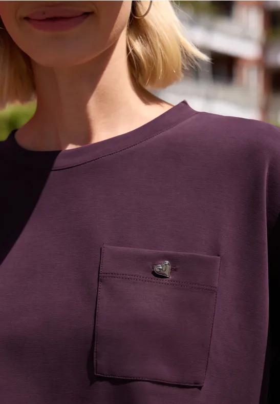 Frau in einem dunkellila Shirt mit aufgesetzter Brusttasche und kleinem goldenen Emblem.