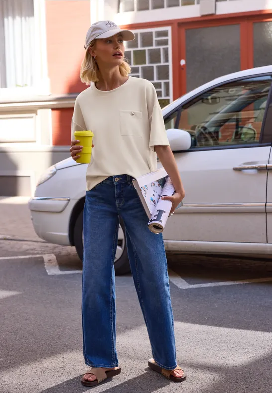 Eine Person in T-Shirt, weiten Jeans und Flip-Flops läuft mit Kaffee und Zeitung eine Straße entlang.