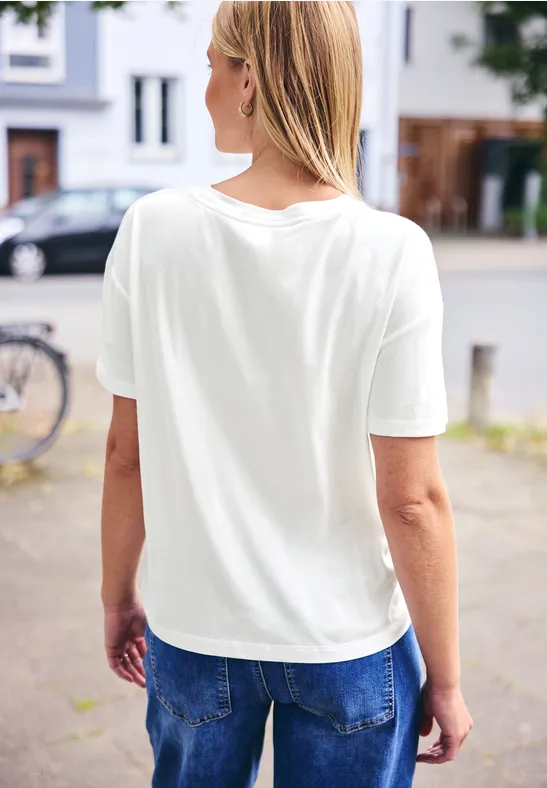 Frau in weißem T-Shirt und blauen Jeans, von hinten stehend auf einem Gehweg neben einem Fahrrad.