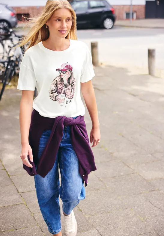 Ein junges blondes Mädchen spaziert in Jeans, T-Shirt mit Hunde-Motiv und Sweatshirt um die Taille.