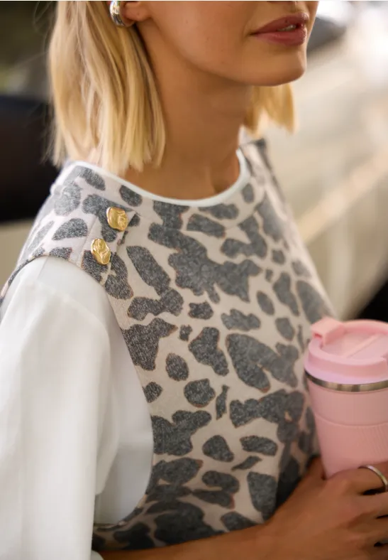 Frau trägt graues Leo-Stricktop über weißem Shirt, mit goldenen Schulter-Pins und hält rosa Kaffeebecher.