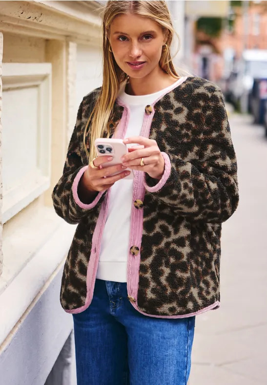 Frau lehnt in Leopardenjacke an Hauswand und schreibt auf ihr Smartphone.