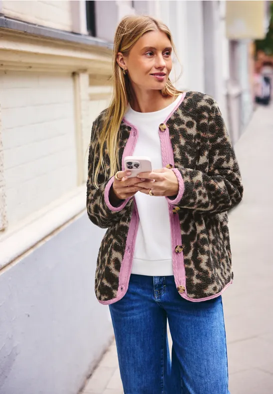 Junge Frau in Jeans und Leopardenstrickjacke hält einen Kaffee to go auf der Straße.