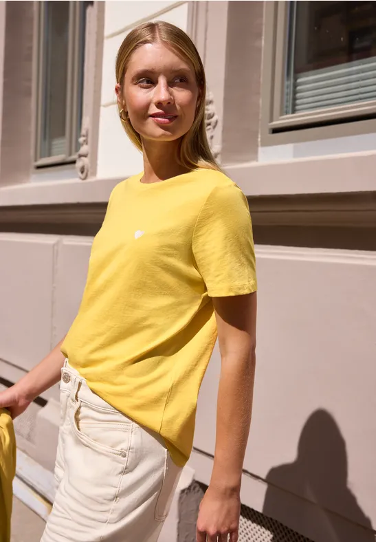 Femme en t-shirt jaune et pantalon clair devant une façade, regard tourné de côté.