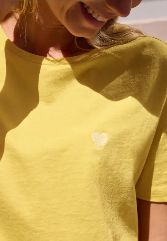 Gros plan sur une femme en t-shirt jaune avec un petit cœur blanc sur la poitrine.