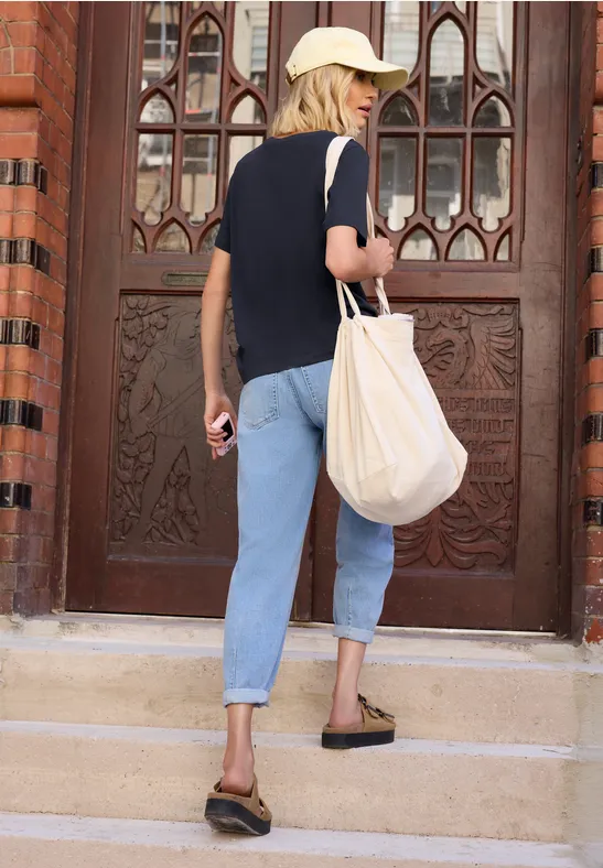 Eine Frau in Jeans, T-Shirt, Cap und Sandalen mit Stofftasche besteigt Stufen vor einer verzierten Holztür.