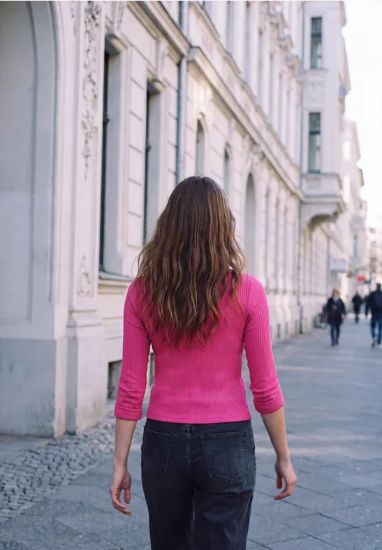 Eine Frau in pinkem Oberteil und Jeans geht eine breite Straße mit historischen Gebäuden entlang.
