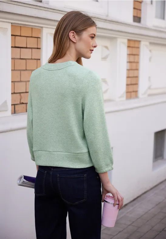 Frau im mintgrünen Pullover und dunklen Jeans steht seitlich vor weißer Hausfassade und hält rosa Trinkbecher.