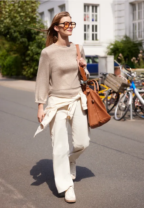 Eine junge Frau in hellem Pullover, weißen Hosen und Sonnenbrille schlendert mit Tasche an sonniger Straße entlang.