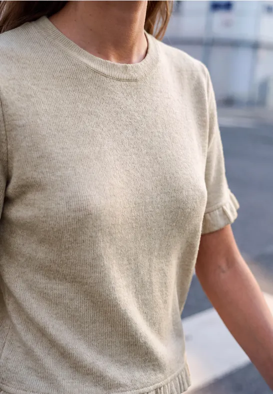 Person im beigen T-Shirt mit aufgekrempelten Ärmeln, im Hintergrund unscharfe städtische Umgebung.