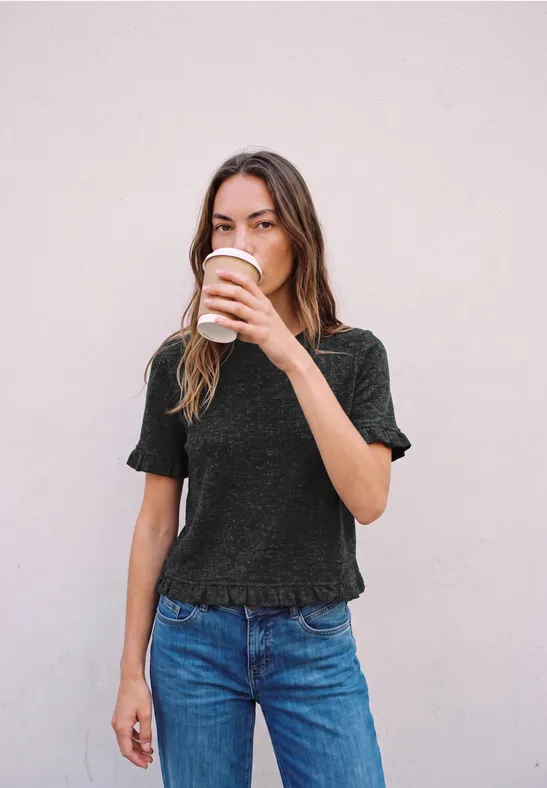 Eine junge Frau trinkt aus einem Pappbecher Kaffee vor einer hellen Wand.