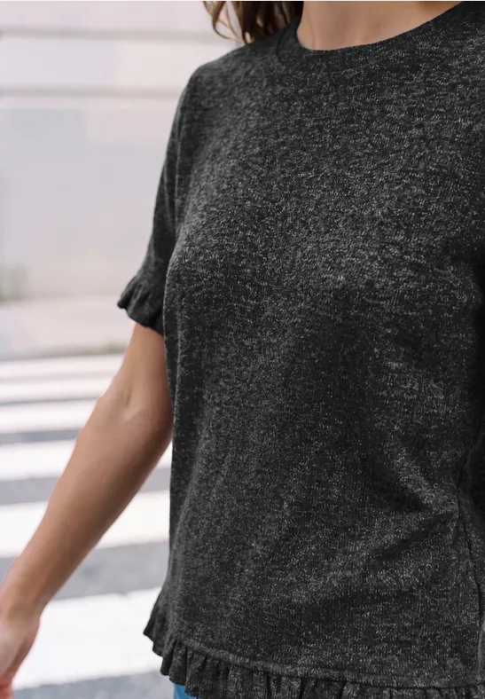 Frau trägt dunkelgraues, leicht glitzerndes T-Shirt mit kurzen Rüschenärmeln und Rüschenbund.