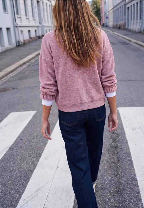 Eine Frau in rosa Pullover und dunklen Jeans überquert von hinten einen Zebrastreifen in der Stadt.