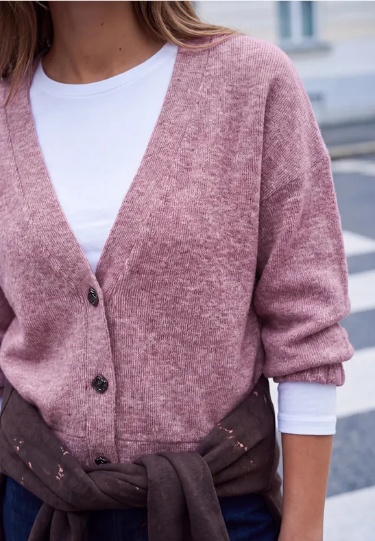 Eine Person trägt eine rosafarbene Strickjacke mit schwarzen Knöpfen und einen braunen Pullover um die Taille.