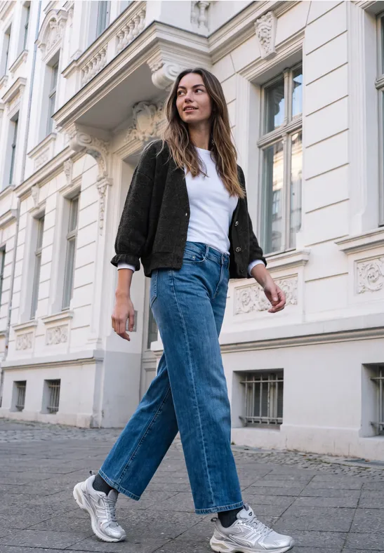 Junge Frau in Jeans, weißem Shirt und silbernen Sneakern spaziert vor einem klassischen Altbau.