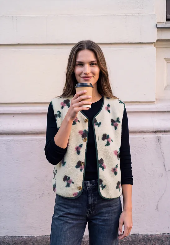 Jeune femme devant un mur, souriante, tenant un café à emporter, vêtue d’une veste à motifs, pull noir et jean.