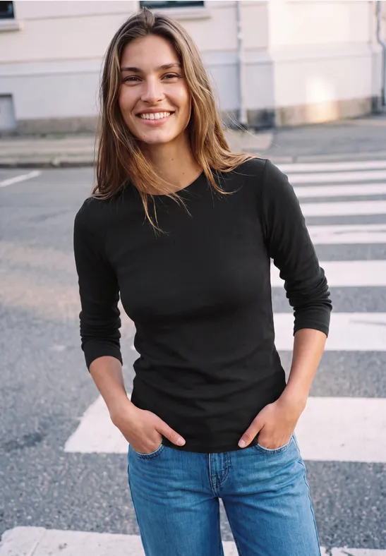 Eine lächelnde junge Frau in schwarzem Oberteil und Jeans steht auf einem Zebrastreifen.