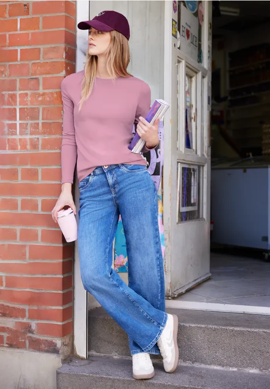 Junge Frau in violettem Langarmshirt, Jeans und Cap lehnt an Backsteinwand und hält Bücher sowie einen Becher.