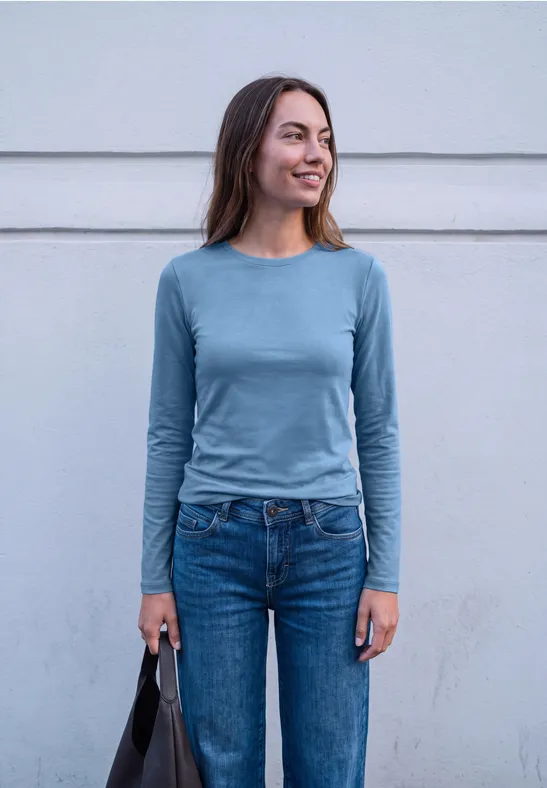 Eine lächelnde junge Frau in blauem Shirt und Jeans mit Tasche in der Hand.