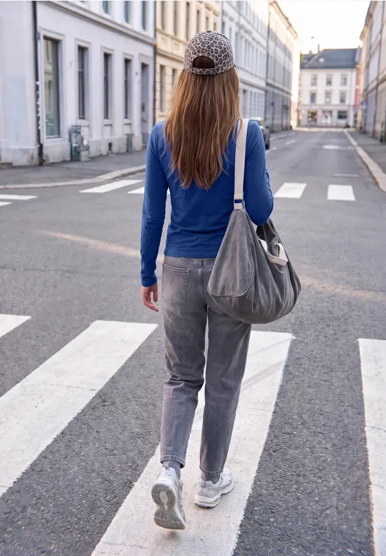 Frau in blauem Oberteil und grauer Hose mit Tasche läuft über einen Zebrastreifen in einer ruhigen Stadtstraße.