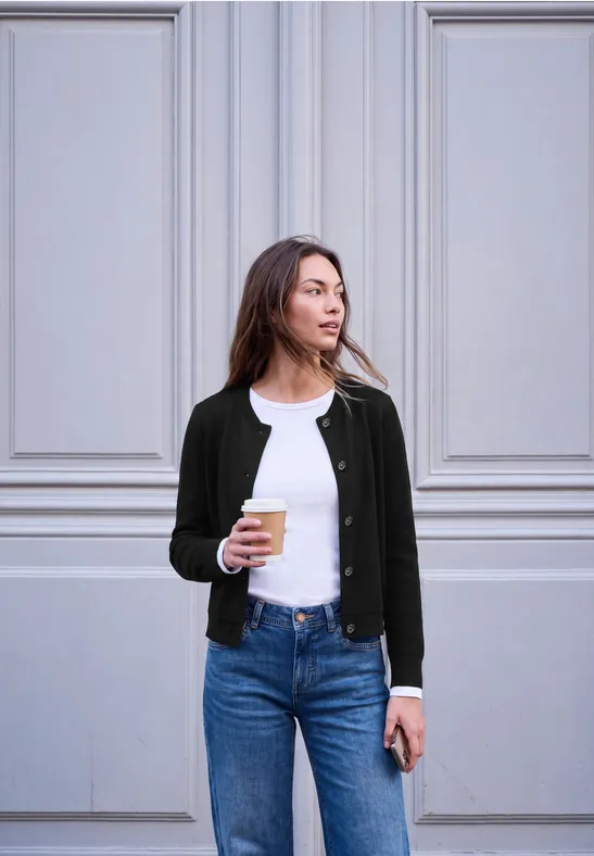 Een vrouw staat voor een lichte muur, draagt een spijkerbroek, t-shirt en vest en houdt koffie en smartphone vast.