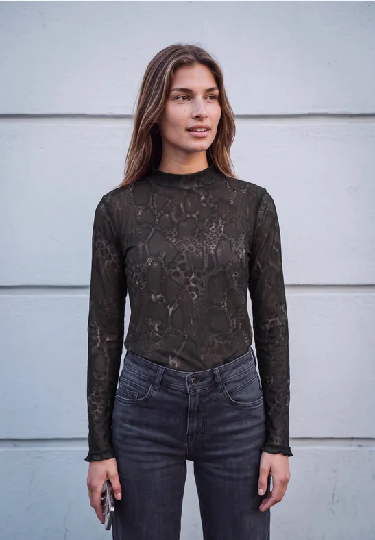 Vrouw met bruin haar voor een grijze muur draagt een donker kanten top en spijkerbroek, houdt een smartphone vast.