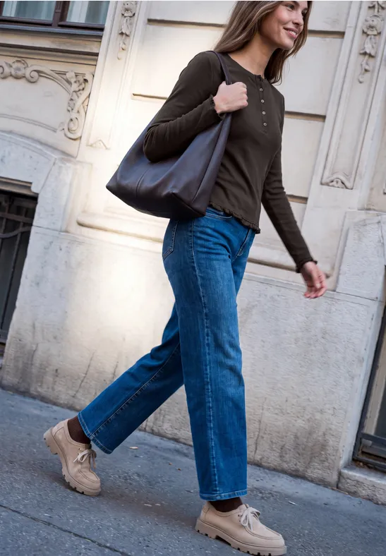 Eine Frau in Jeans, Sneakern und brauner Bluse trägt eine Tasche und läuft an einer Hausfassade vorbei.