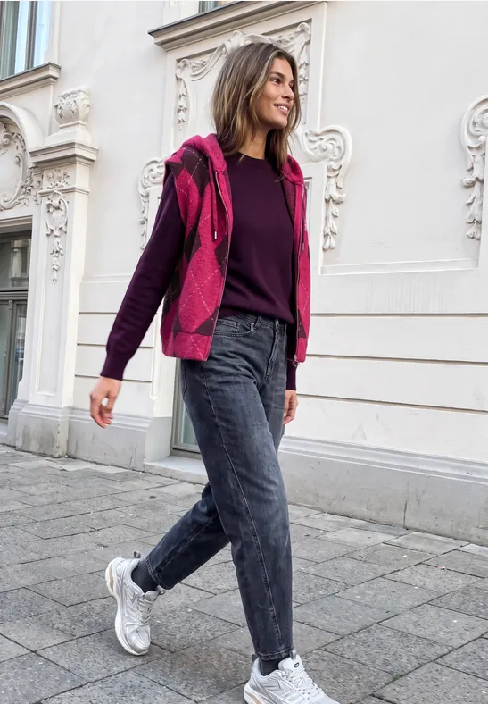 Eine Frau in rosa Jacke und Jeans beim Spaziergang vor einer Gebäudefassade.