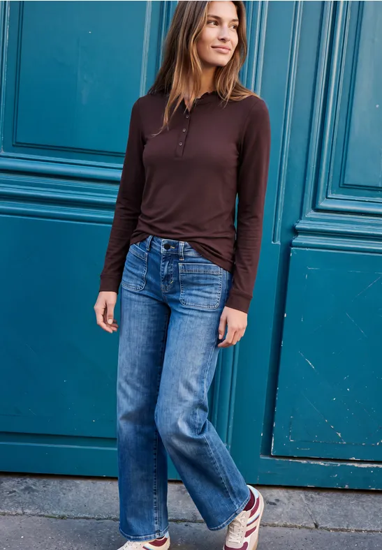 Frau steht vor einer blauen Tür, trägt braunes Oberteil, Jeans und Sneaker, lächelt und schaut zur Seite.